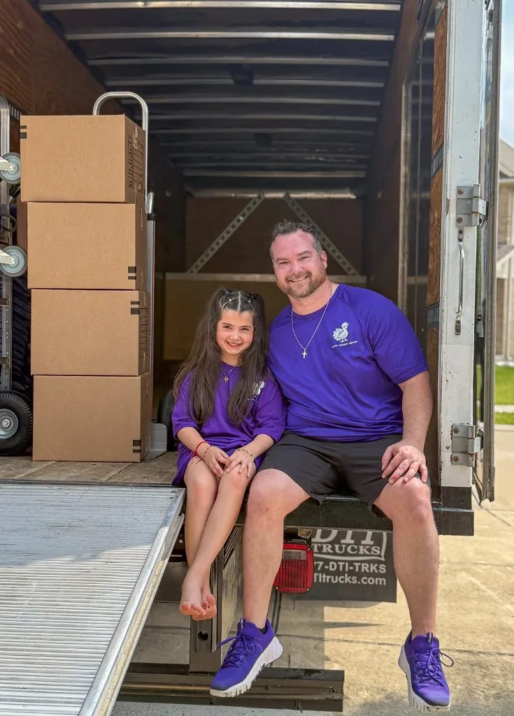 A Purple Peacock mover and a young helper in purple sitting on the lift gate of a loaded moving truck